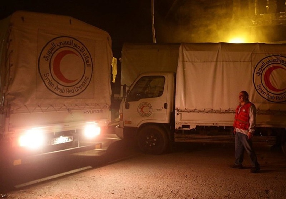 Syrian Arab Red Crescent trucks with distinctive Red Crescent logos operating at night, likely delivering humanitarian aid. A worker in a Red Crescent vest is visible walking near the vehicles. The image shows the logistics aspect of humanitarian operations with vehicles serving as basic transportation technology for aid delivery.